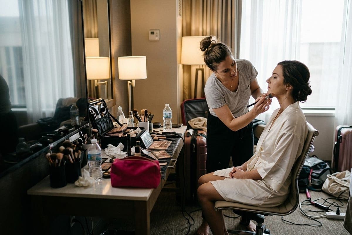 A makeup artist brushing foundation along a client's jawline in a hotel room, their kit spread across the dressing table.